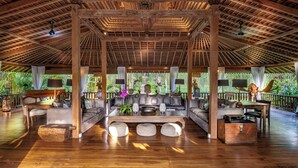 Un vaste espace intérieur doté d’un haut plafond en bois, d’un coin salon central avec des canapés et une table basse, et offrant une vue sur une végétation luxuriante à l’extérieur.
