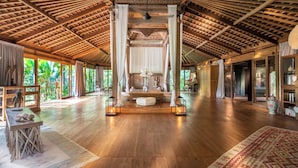 Une pièce spacieuse avec un haut plafond en bois, un espace salon central avec une table, et de grandes fenêtres offrant une vue sur la verdure environnante.