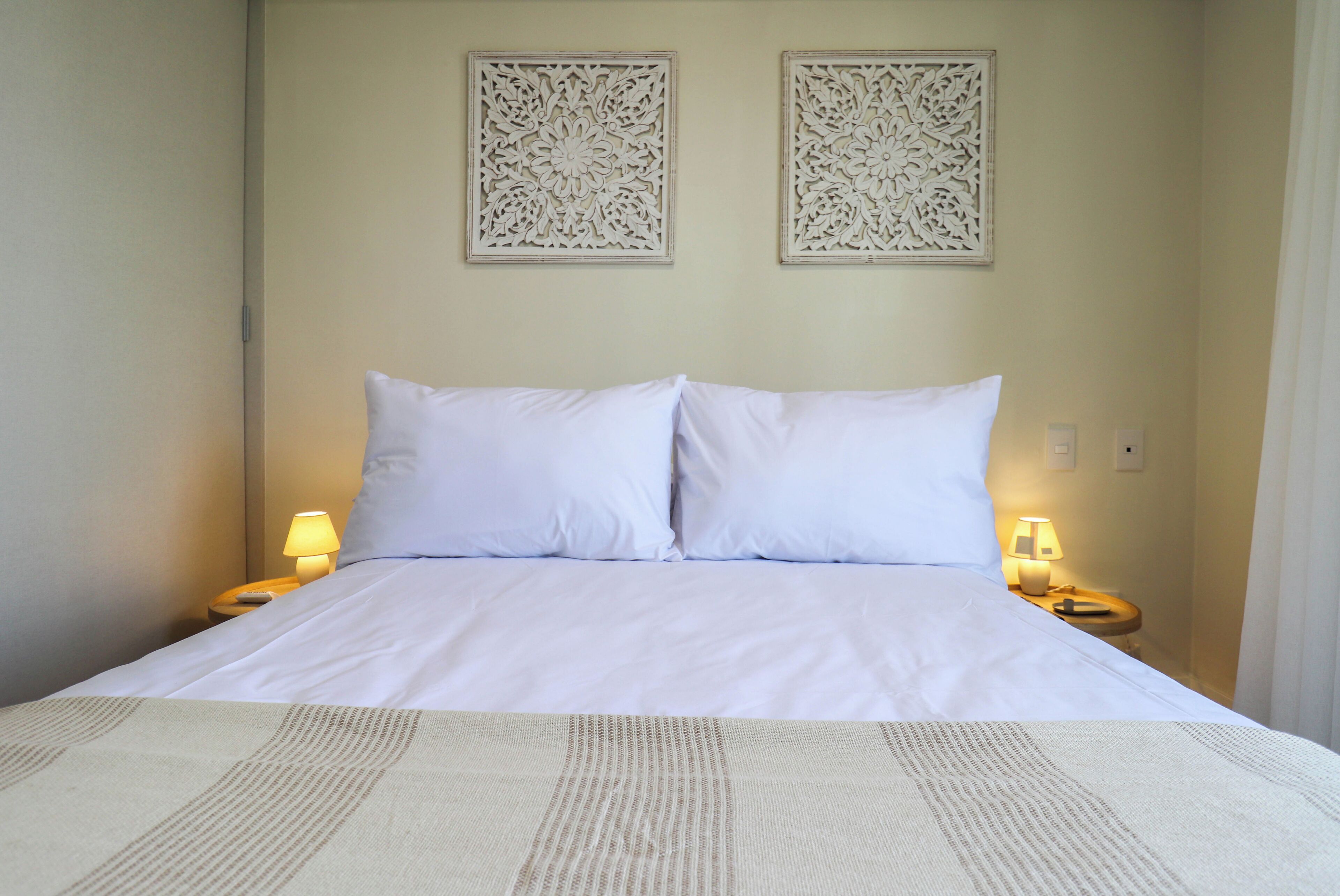 Cama bem arrumada com lençóis brancos, dois painéis decorativos fixados na parede e luminárias de cabeceira de cada lado.