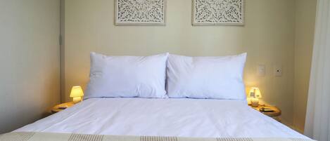 Cama bem arrumada com lençóis brancos, dois painéis decorativos fixados na parede e luminárias de cabeceira de cada lado.