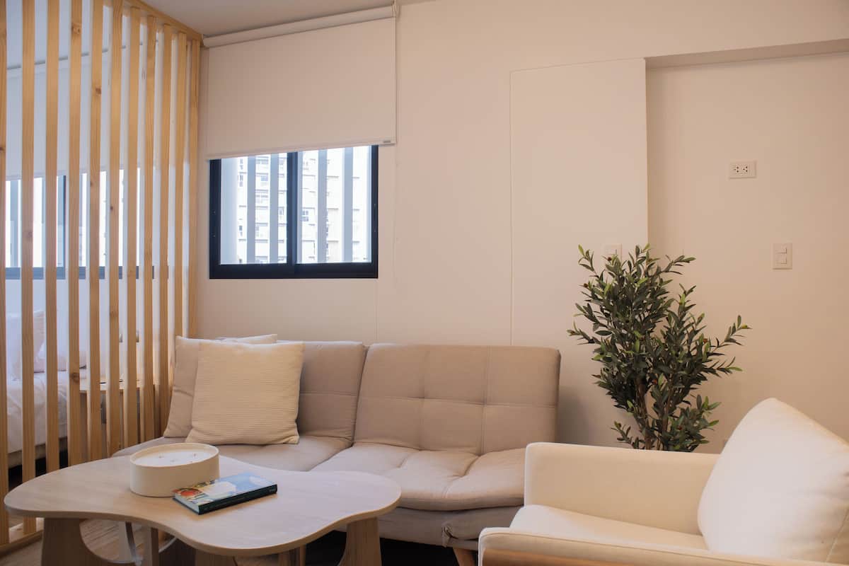 A modern living room with a beige sofa, a small round table with a book, a potted plant, and a window with blinds.