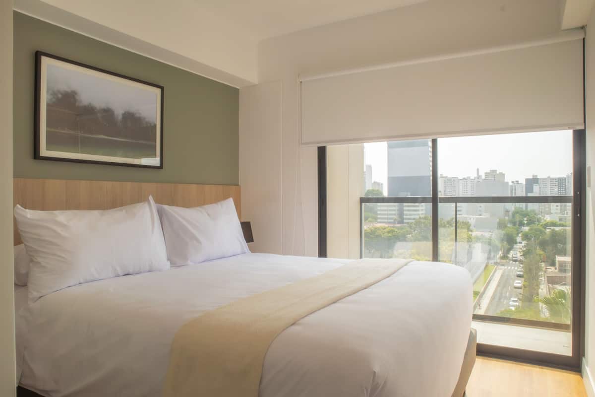 A large bed with white linens in a modern hotel room with a city view.