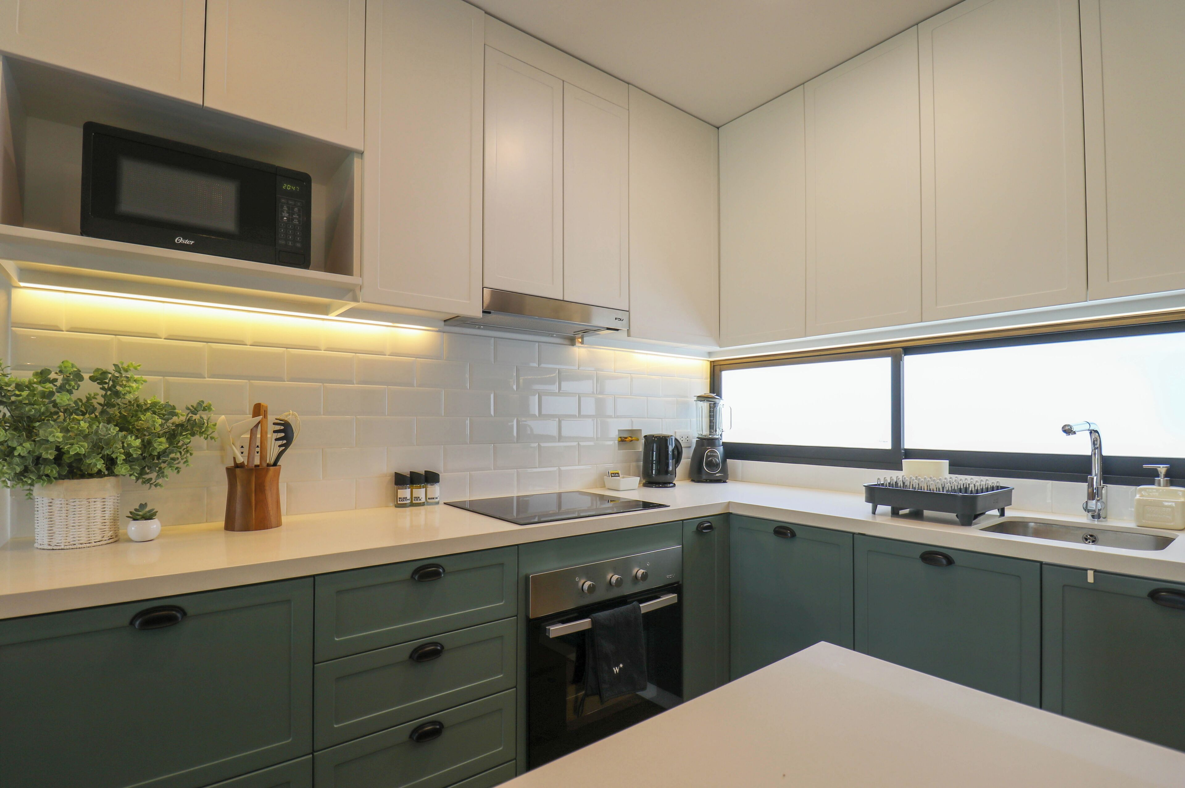Una cocina moderna con armarios blancos, microondas, estufa y lavamanos. Hay una ventana que proporciona luz natural y una planta en maceta sobre la encimera.