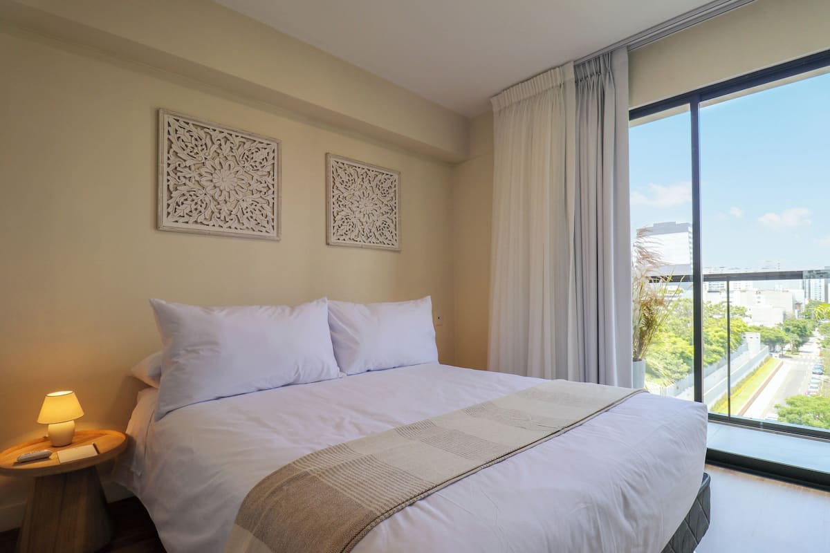 A hotel room with a large bed, a nightstand with a lamp, two wall art pieces, and a large window with a city view.