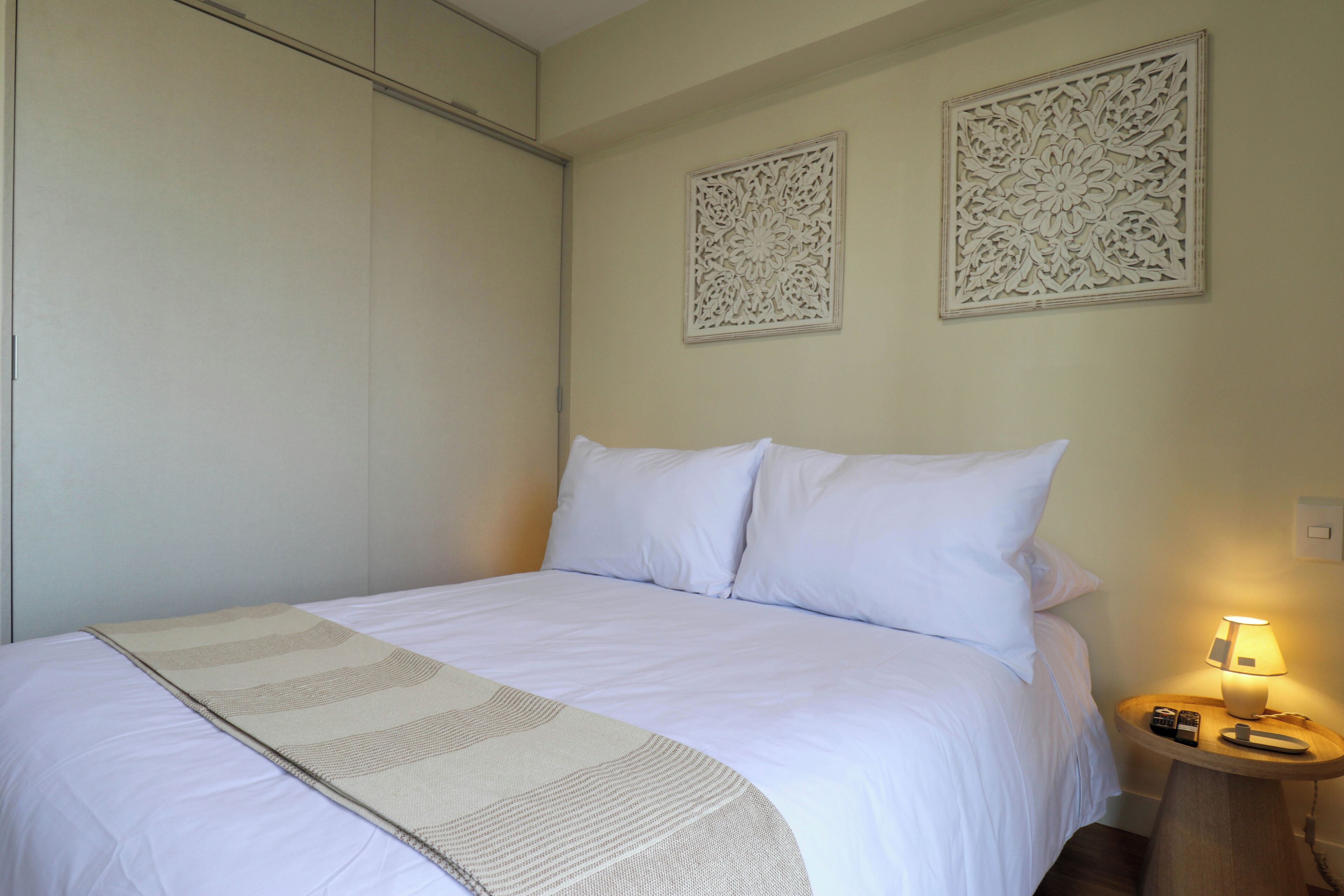 Una cama bien hecha con sábanas blancas y un plaid beige, una lámpara de noche y dos paneles decorativos en la pared.