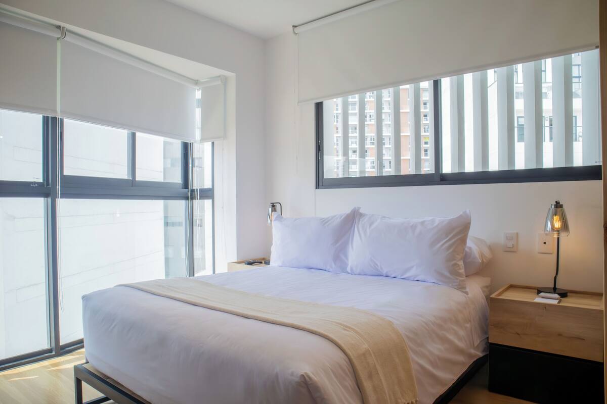A modern hotel room with a large bed, a nightstand with a lamp, and a window with a view of high-rise buildings.