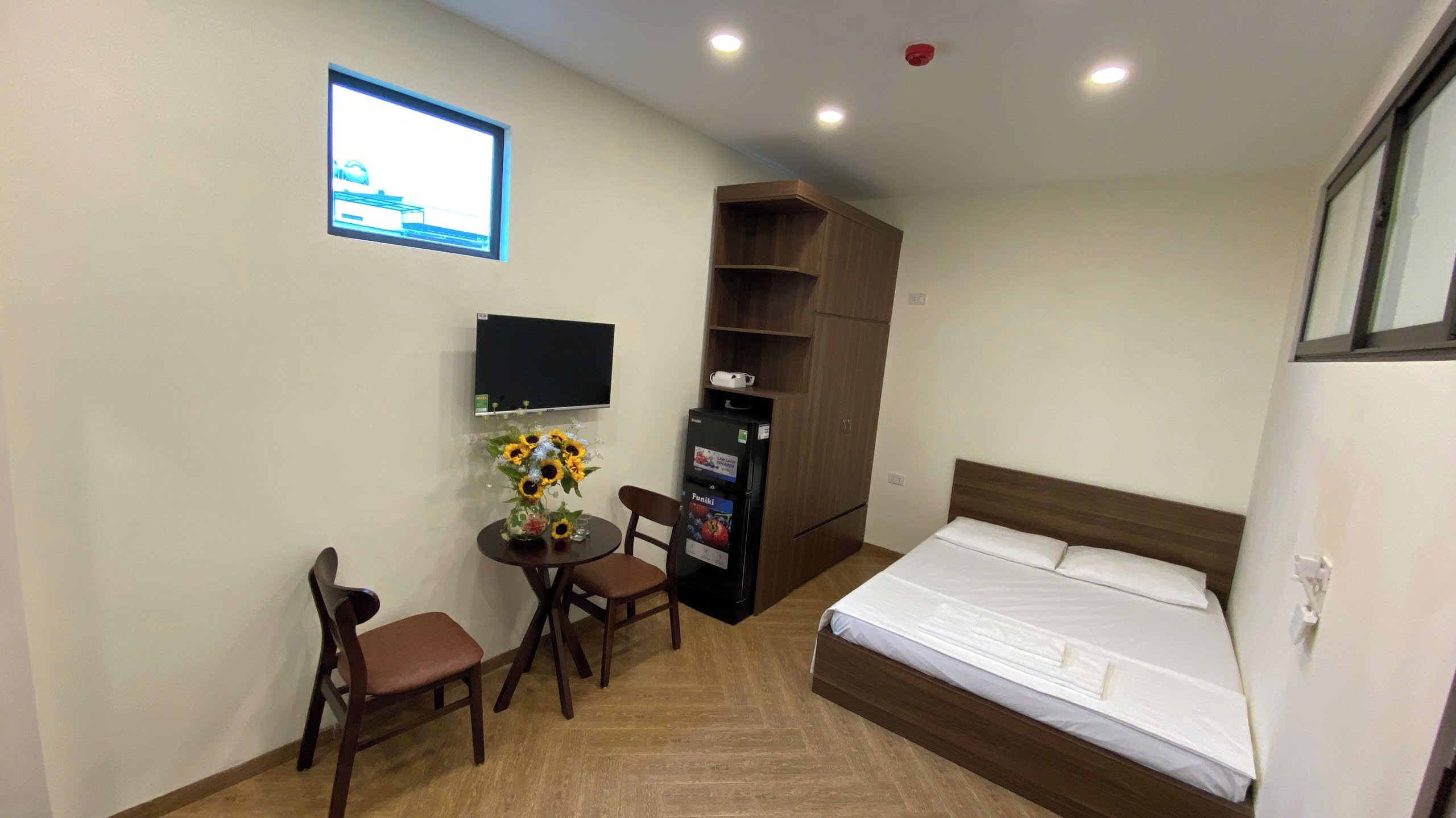 A hotel room with a bed, a desk with a chair, a small table with a vase of flowers, a television mounted on the wall, and a minibar.