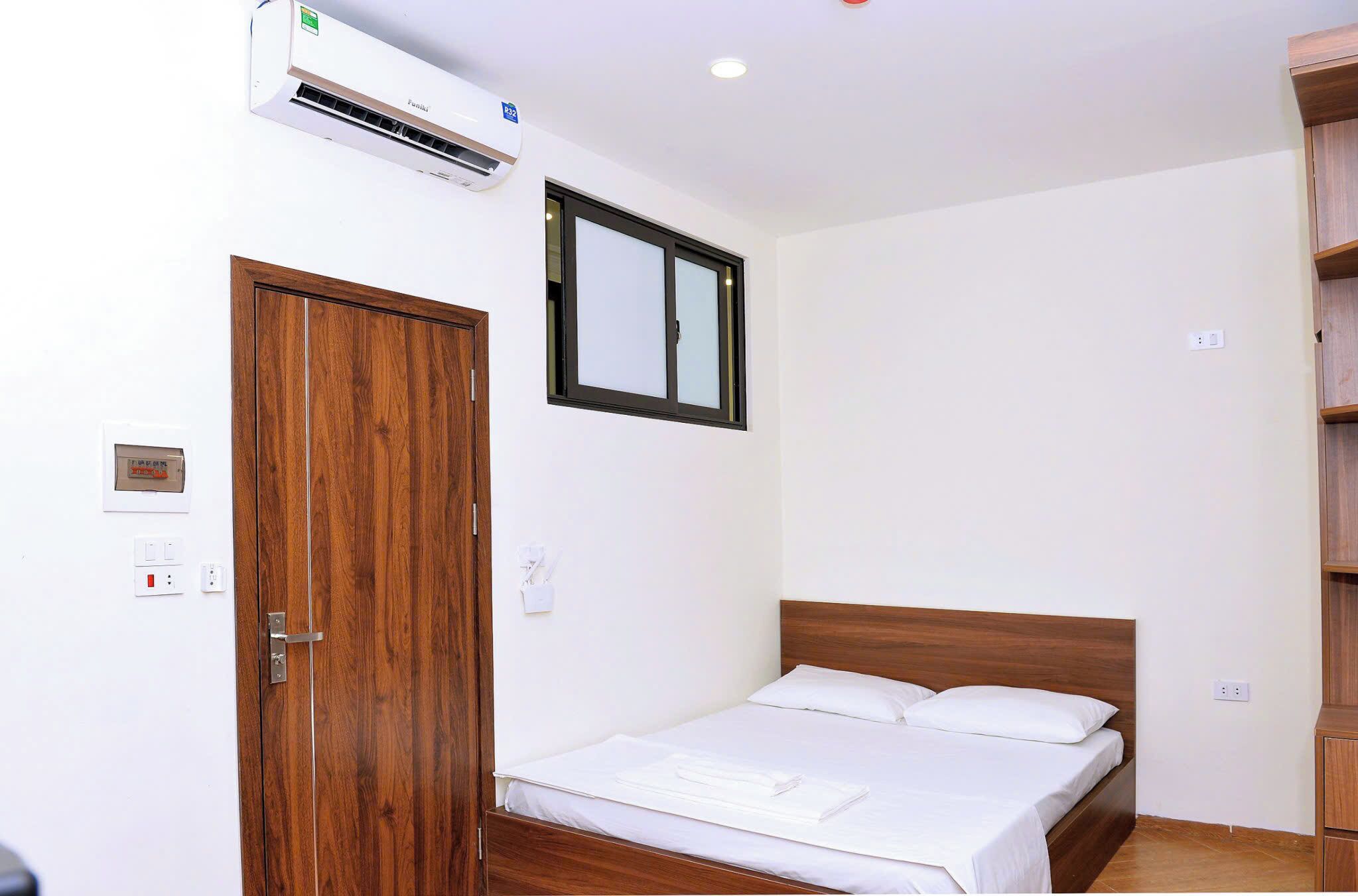 Une chambre d’hôtel avec une porte en bois, un lit avec du linge de maison blanc, un appareil de climatisation, une fenêtre et un thermostat fixé au mur.