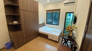 Una habitación pequeña y ordenada con una cama, un armario de madera, una mesita con un florero y una ventana con vistas a la vegetación.