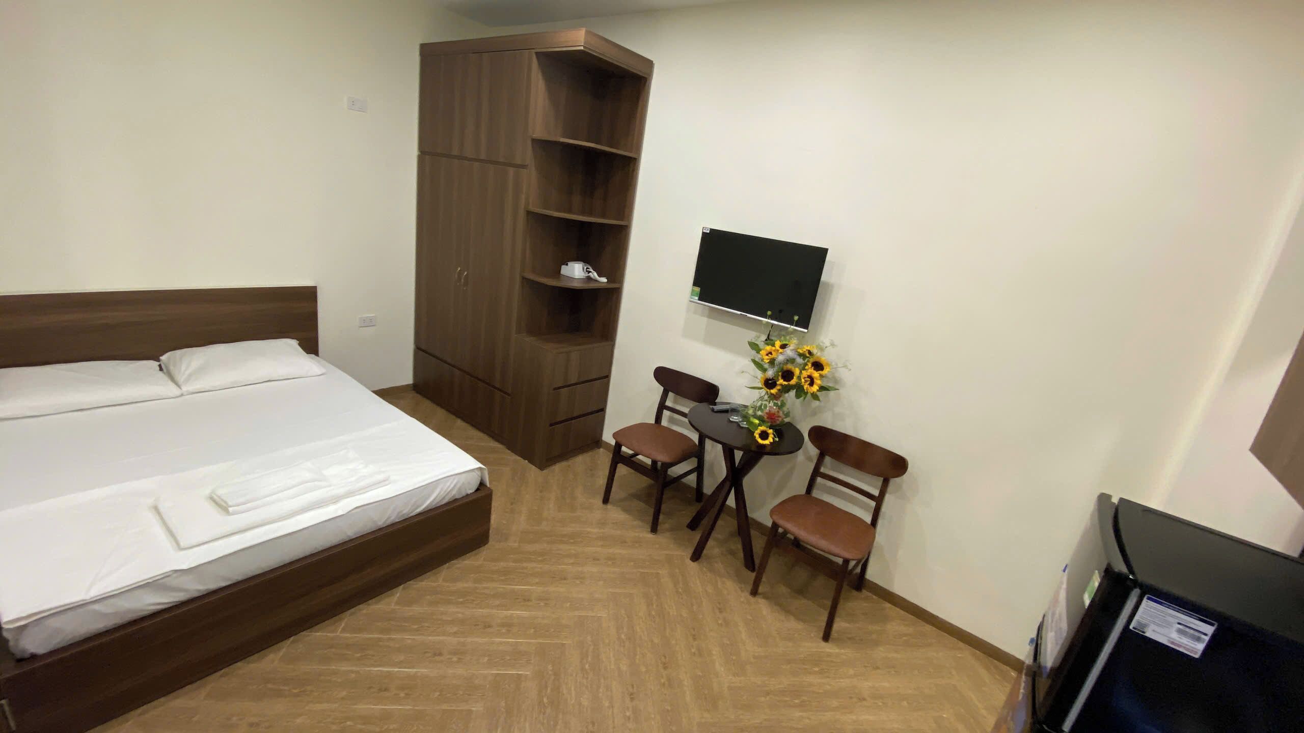 Ein Hotelzimmer mit einem großen Bett, einem Holzschrank, einem Schreibtisch mit zwei Stühlen, einem an der Wand montierten Fernseher und einer Vase mit Sonnenblumen.