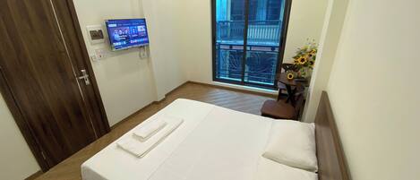 Habitación de hotel con una cama, un televisor montado en la pared, una ventana con vistas a un balcón y una pequeña mesa con un jarrón de flores.