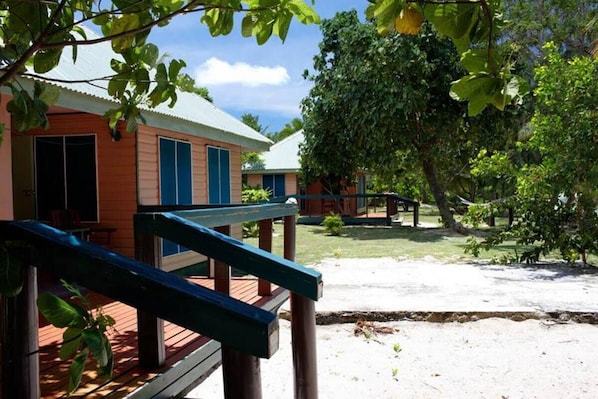A wooden deck with a blue railing, a building with a red facade and blue shutters, and a large tree in the background. - Long Beach Escape (Matathawa Island)