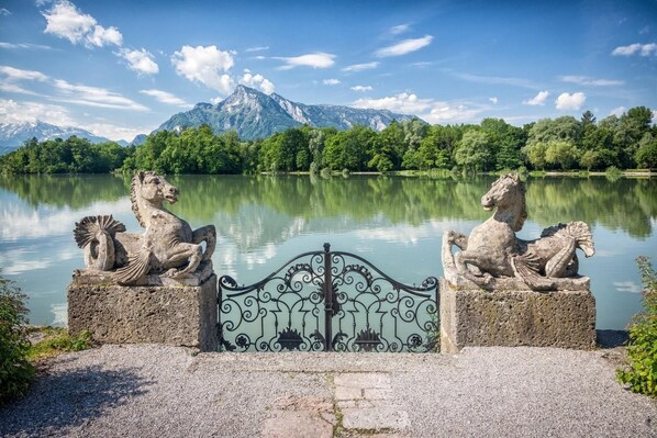 Une statue de lion en pierre, posée sur un piédestal, au bord d’un lac, avec des montagnes en arrière-plan.