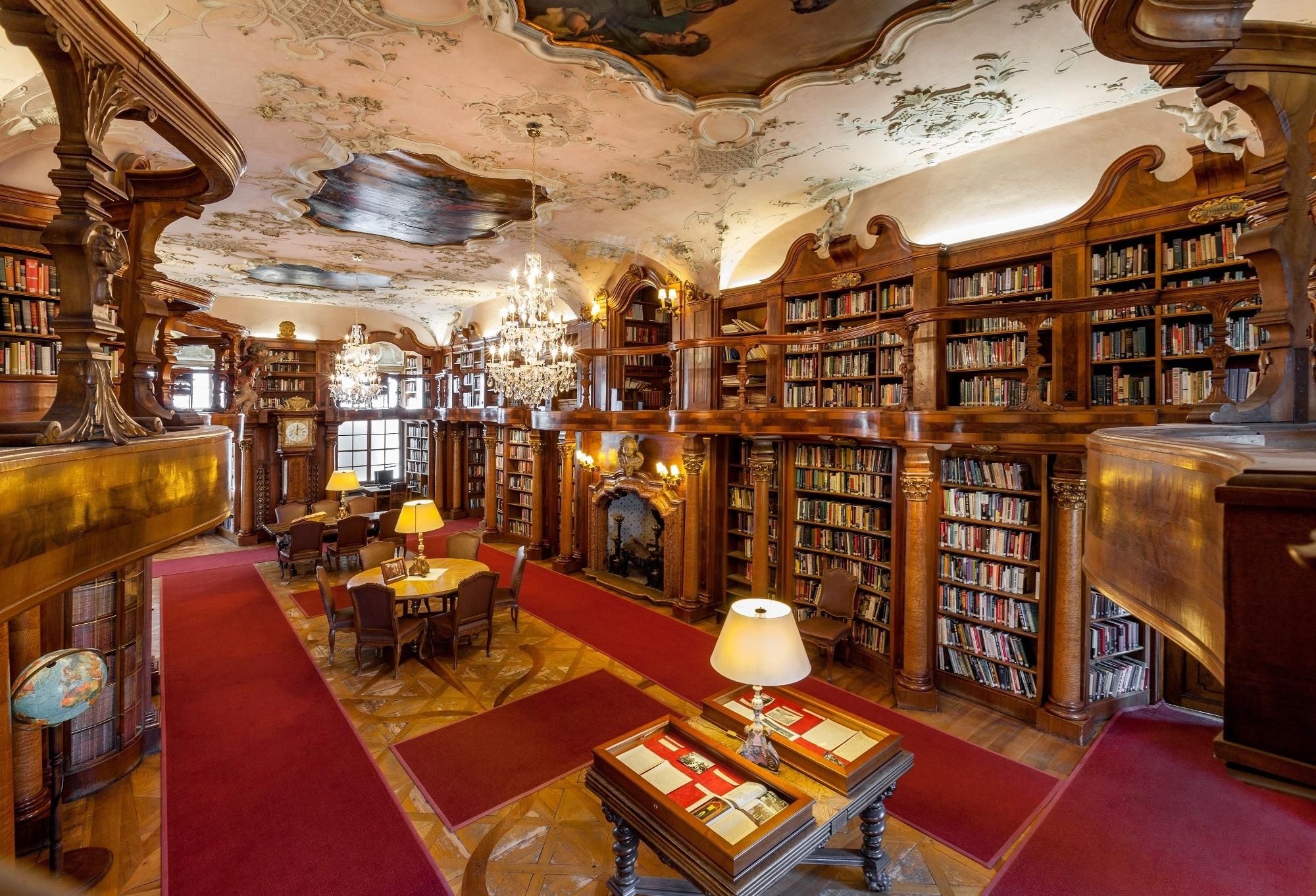 A grand library with ornate woodwork, bookshelves, a fireplace, and a chandelier.