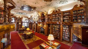 A grand library with ornate woodwork, bookshelves, a fireplace, and a chandelier.