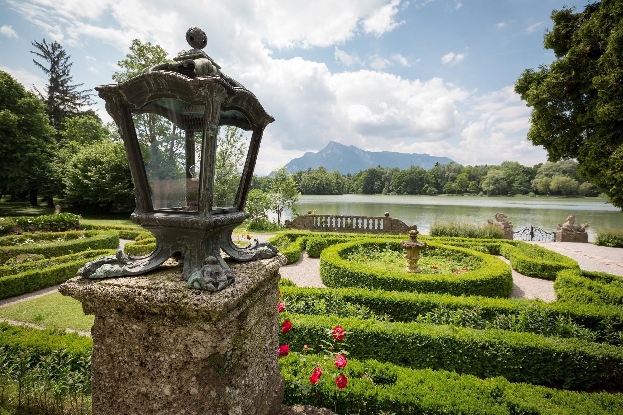 Ein Garten mit einer Steinhlaterne, geschnittenen Hecken und Blick auf einen See.