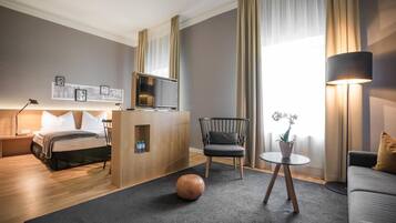 Ein modernes Hotelzimmer mit Bett, Schreibtisch, Fernseher, Stuhl, einem kleinen Tisch mit Pflanze und einer Couch.