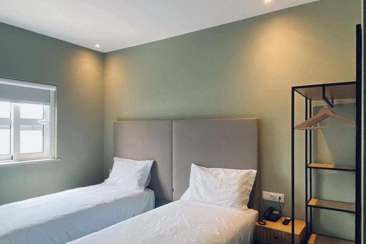 A hotel room with two single beds, a bedside table with a lamp, a wardrobe, and a window with blinds.