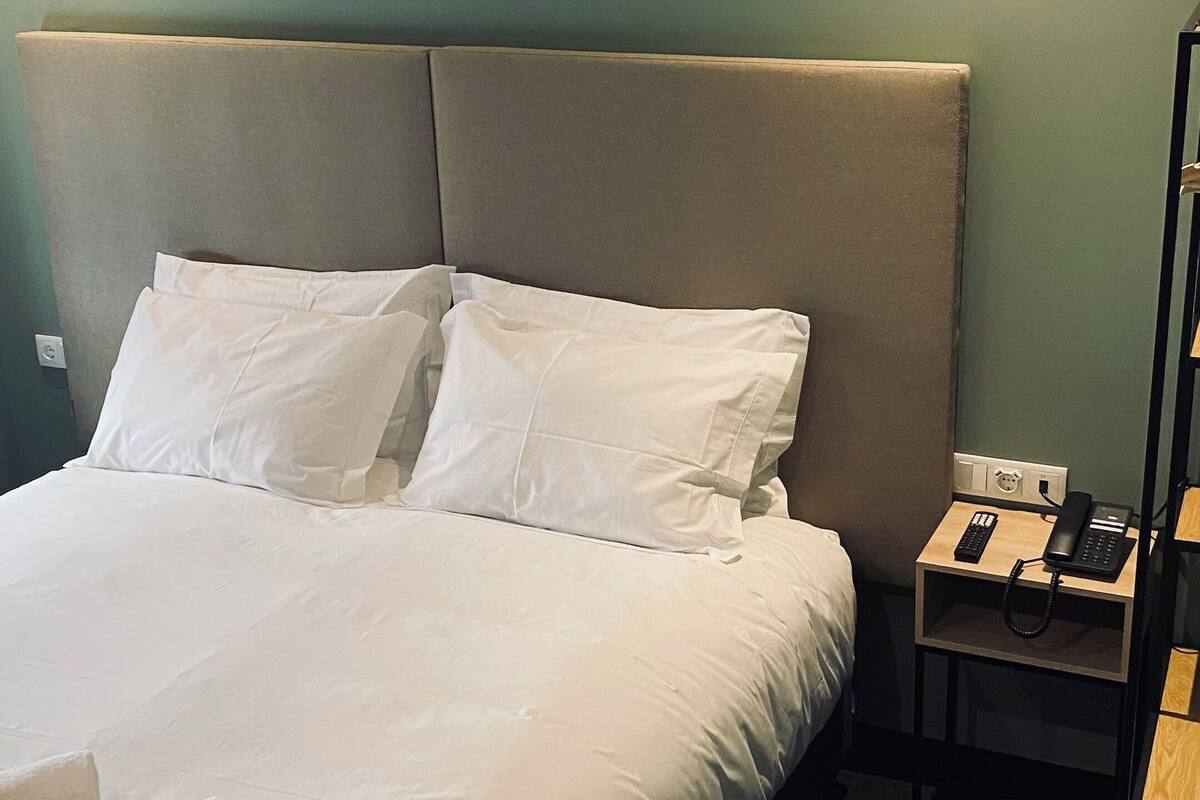 A neatly made bed with white linens and pillows, a bedside table with a phone and a lamp, and a green wall in the background.