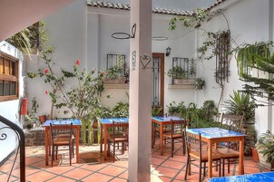 Un patio al aire libre con suelo de baldosas, mesas y sillas de madera, y plantas en macetas.
