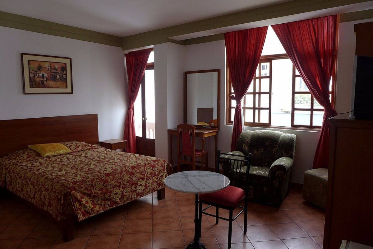Habitación de hotel con cama, una silla, una mesita, un espejo y una ventana con cortinas.
