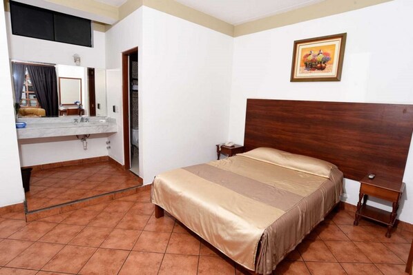 Habitación de hotel con cama, cabecera de madera, mesita de noche, tocador, espejo y baño con lavabo y toallero.