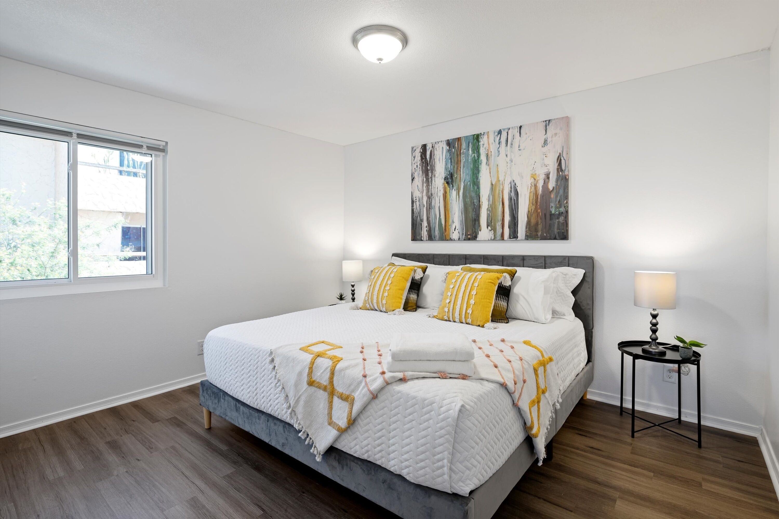 A bedroom with a bed, a nightstand, a window, and a painting on the wall.