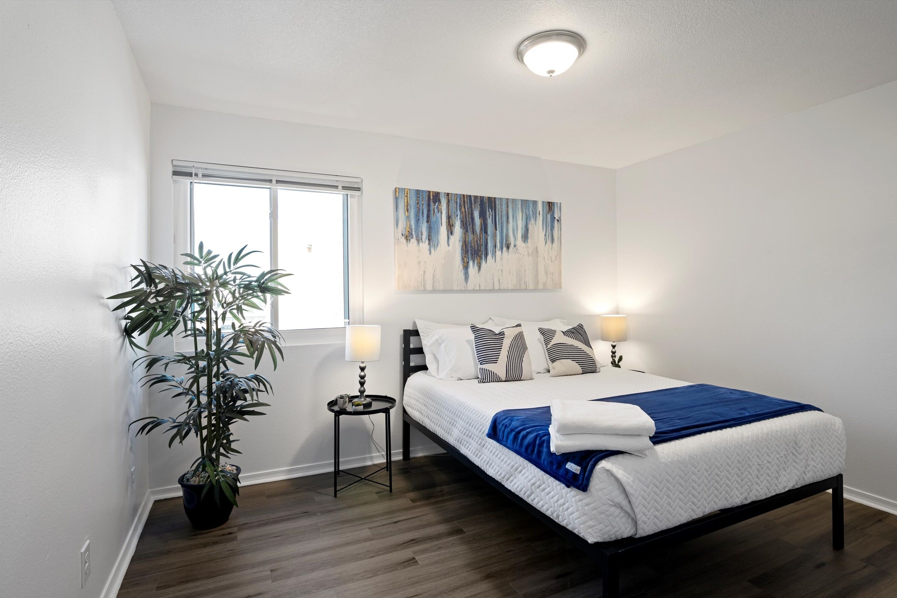 A bedroom with a bed, bedside lamps, a potted plant, and a window.