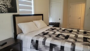 A neatly made bed with a quilt, a bedside table with a remote control, a door, and a wall with a framed picture. - Modern Suite Near Hamilton Airport (Hamilton)