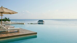 A serene infinity pool with a wooden deck, lounge chairs, and an umbrella, overlooking a calm sea and a distant island structure. - SHA Mexico - Lifestyle Optimization Program - Personalized Meal Plan (Cancun)