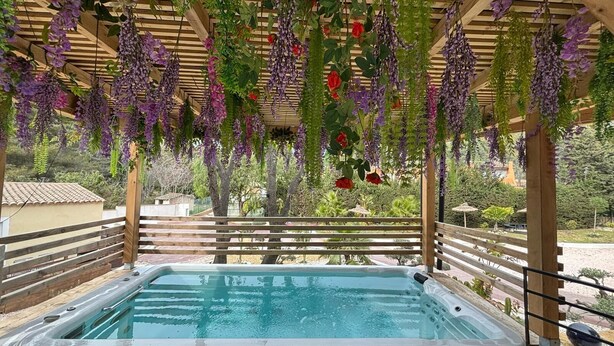 Un bain à remous installé sous une pergola, agrémenté de fleurs violettes en suspension et de roses rouges.