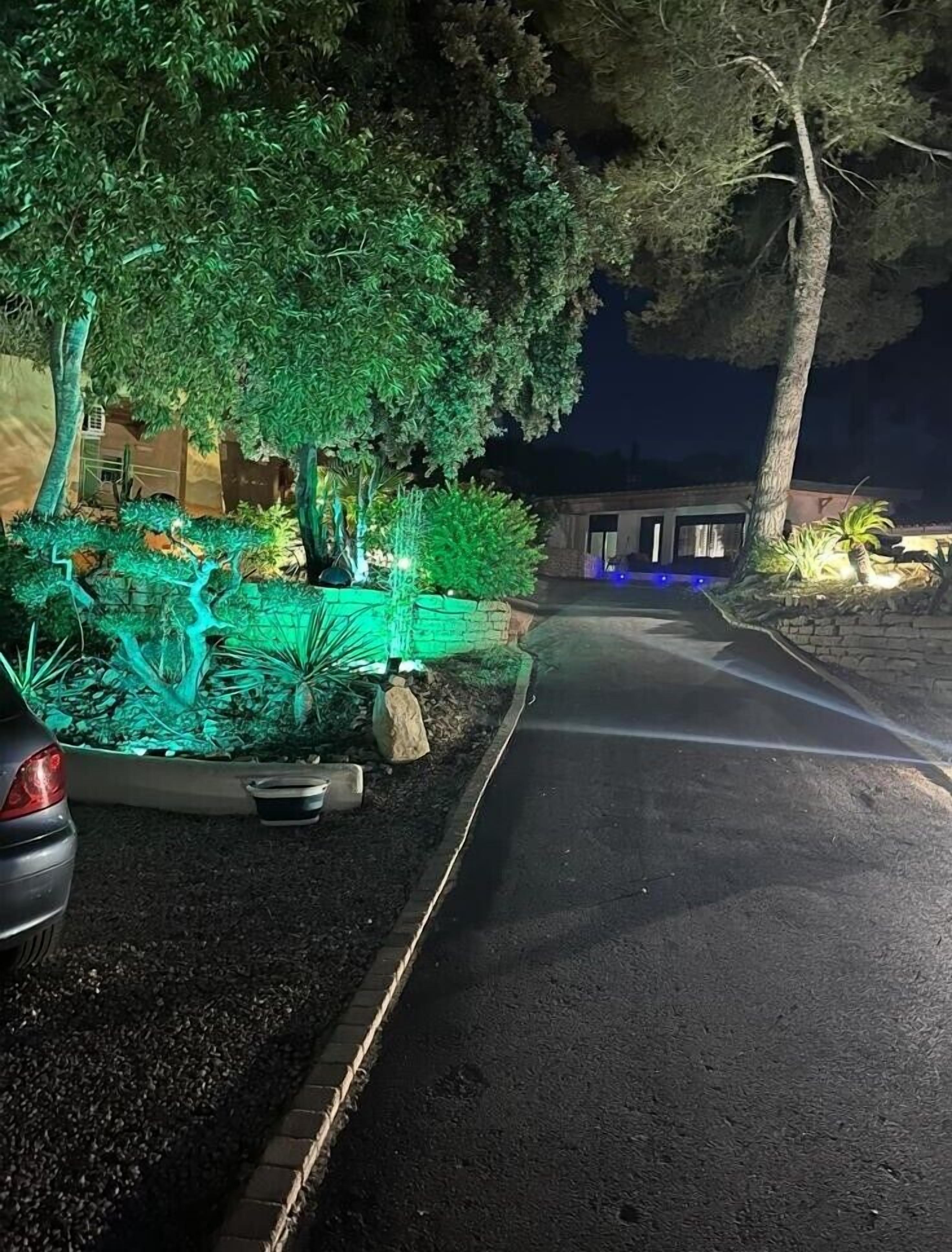 Vista nocturna de una calle con un jardín bien iluminado, un coche estacionado y un edificio al fondo.