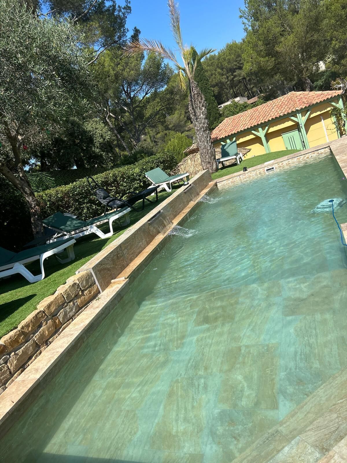 Ein Schwimmbecken mit Steinrand, umgeben von Grünflächen und einem Gebäude mit Ziegeldach.