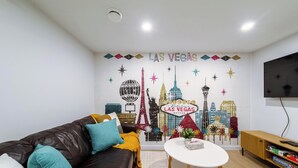 A living room with a cityscape mural, a leather sofa, a small table, and a TV on the wall. - Suite Near WEM: Game Room & Kitchen (Edmonton)