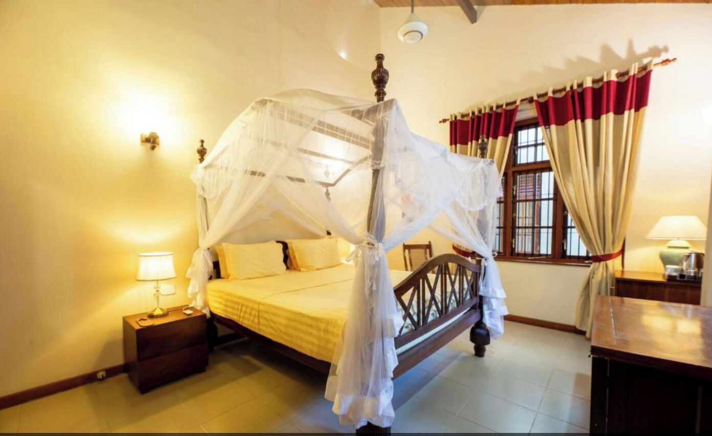 A room with a four-poster bed, a nightstand with a lamp, and a window with curtains.