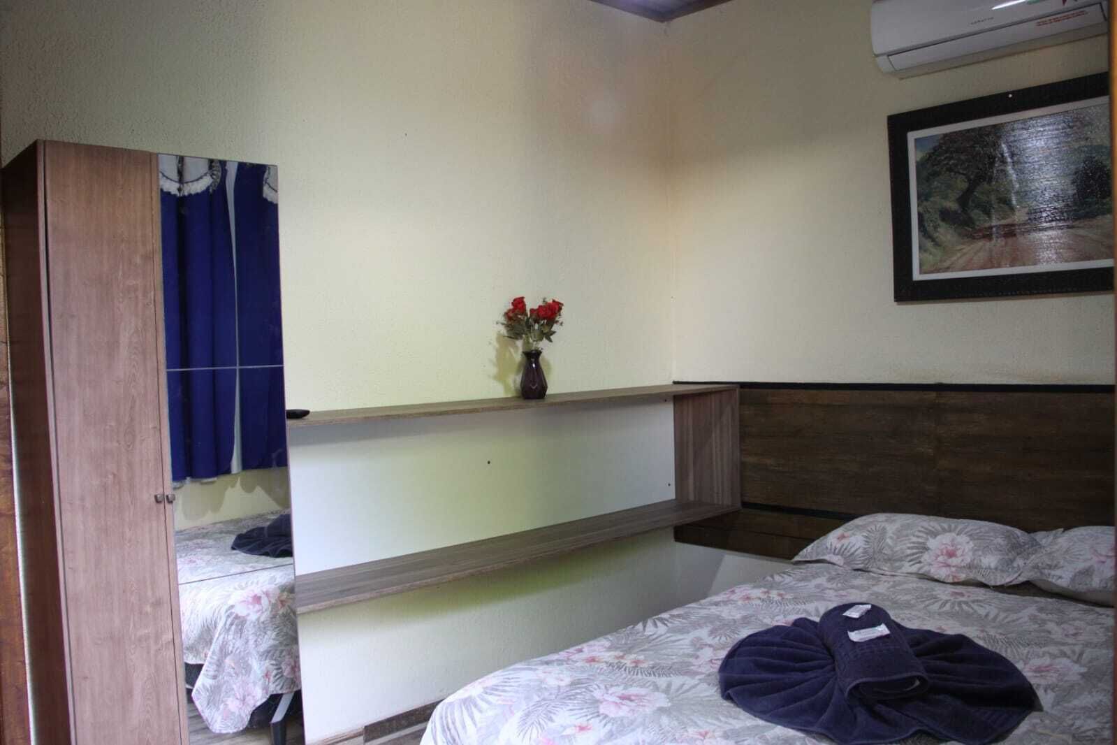 A room with two beds, a wooden wardrobe, a small table with a vase of flowers, and a framed picture on the wall.