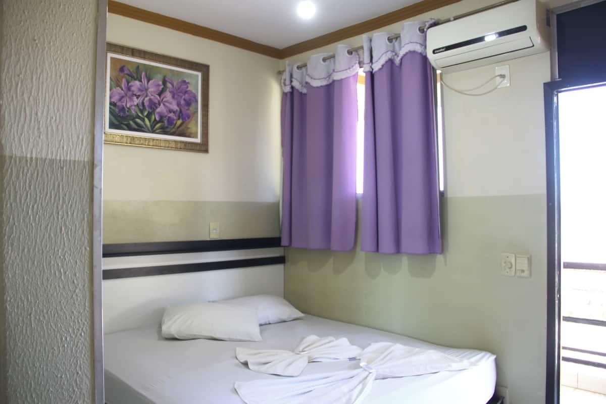 Un lit simple avec du linge de lit blanc, une tête de lit ornée d’une bande horizontale, un climatiseur fixé au mur et une fenêtre avec des rideaux violets.