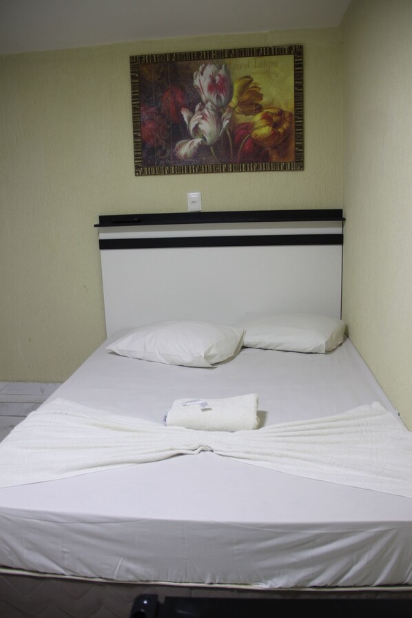 Un lit simple avec une literie blanche, une tête de lit ornée d’une bande horizontale noire et un tableau floral encadré accroché au mur.