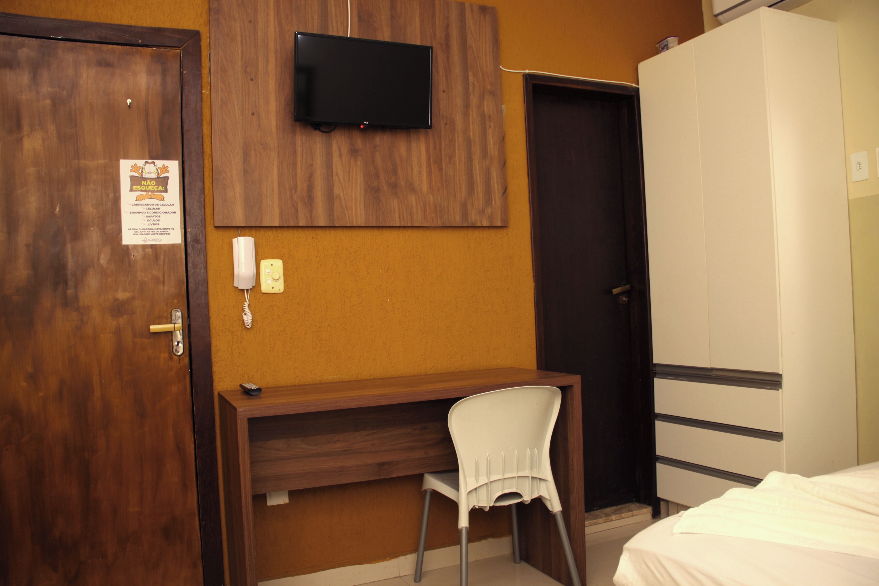 A hotel room with a wooden desk, a white chair, a television mounted on the wall, a wardrobe, and a door.