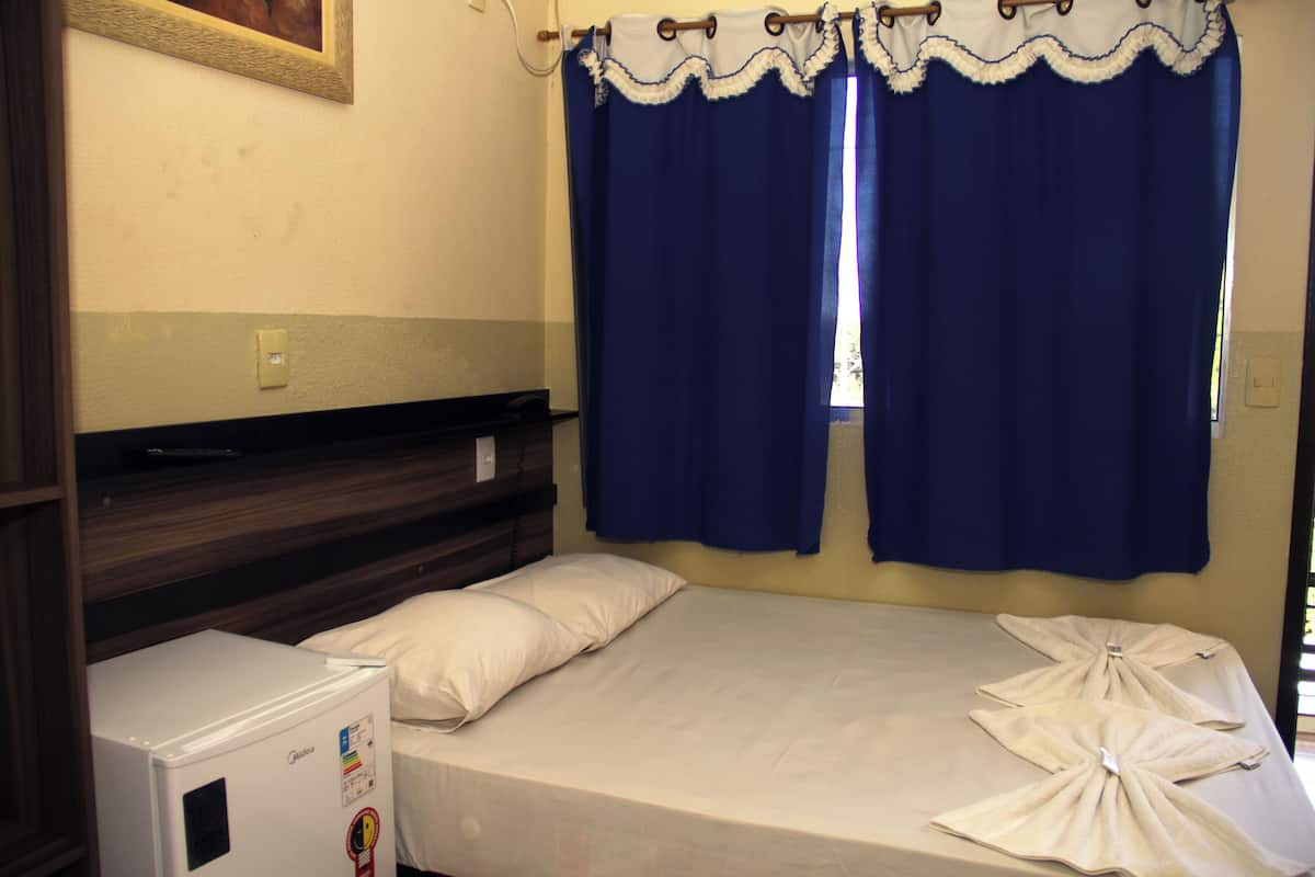 Une petite chambre avec un lit simple, une tête de lit en bois, un petit réfrigérateur et une fenêtre avec des rideaux bleus.