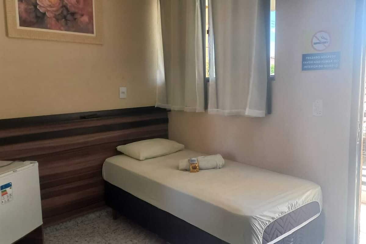 Une petite chambre avec un lit simple, une tête de lit en bois, une table de chevet avec un mini-réfrigérateur, une fenêtre avec des rideaux et un panneau indiquant qu’il est interdit de fumer dans la chambre.