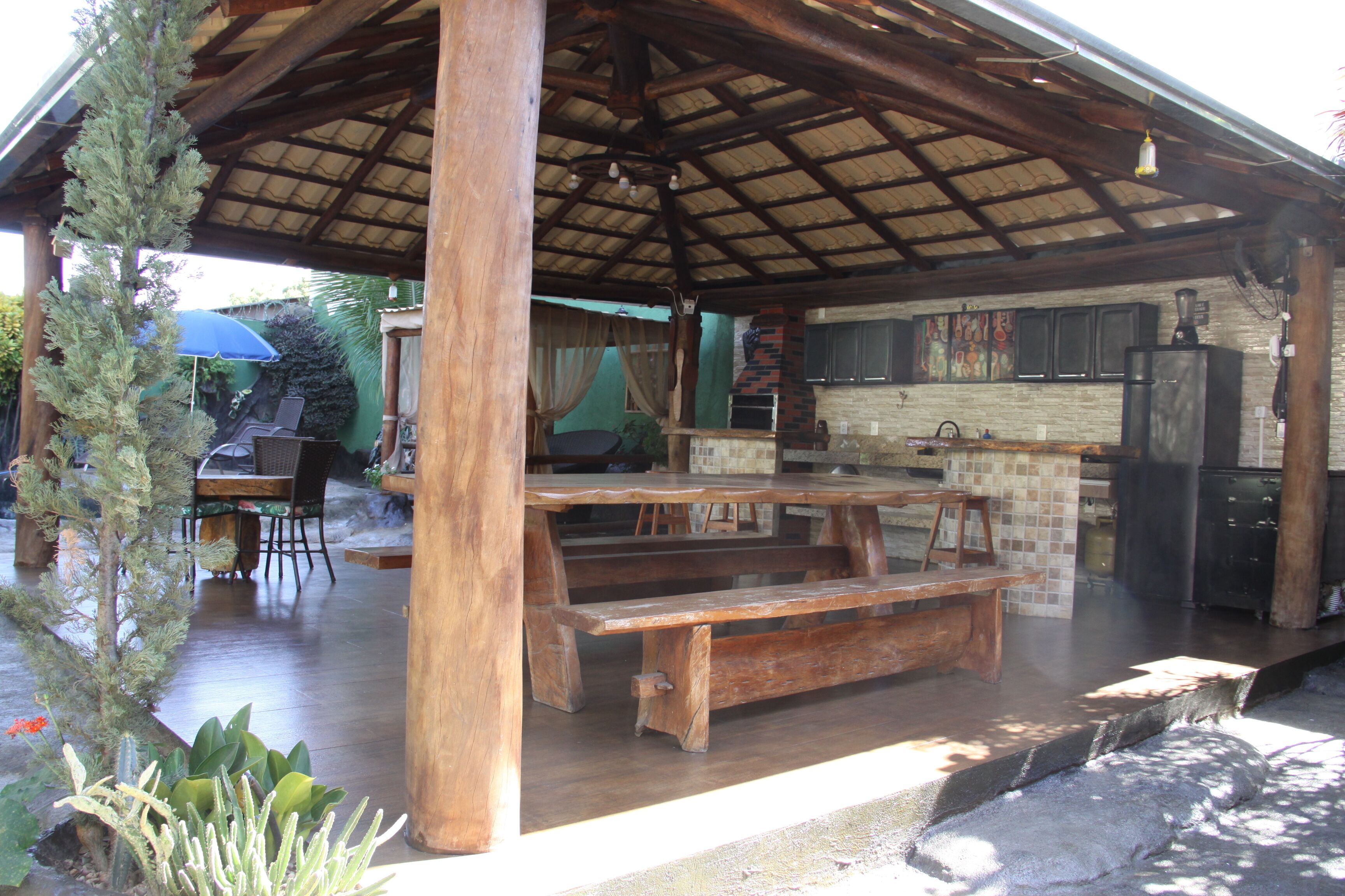 A covered outdoor area with wooden benches, a grill, and a kitchen area with a refrigerator.
