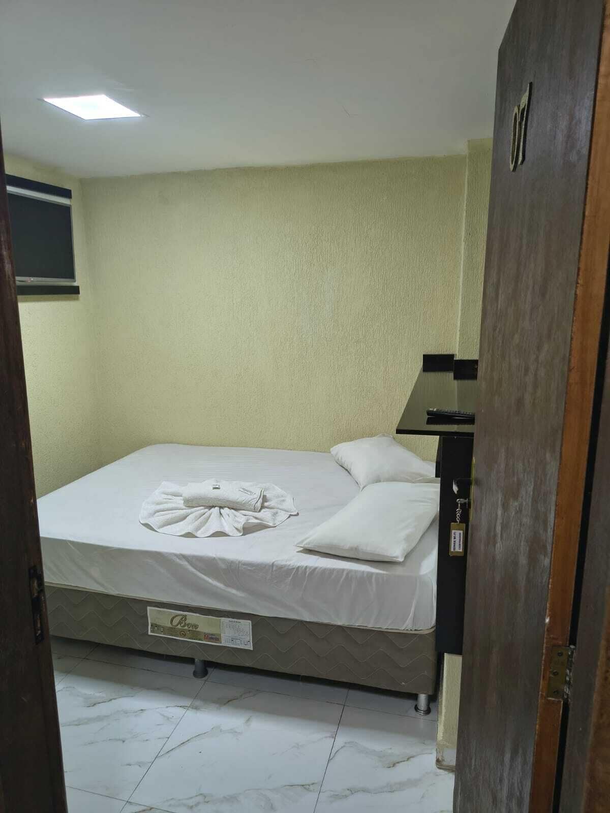 Un petit lit bien fait, avec un matelas et des oreillers blancs, une table de chevet en bois sur laquelle se trouve une lampe, et un sol carrelé.