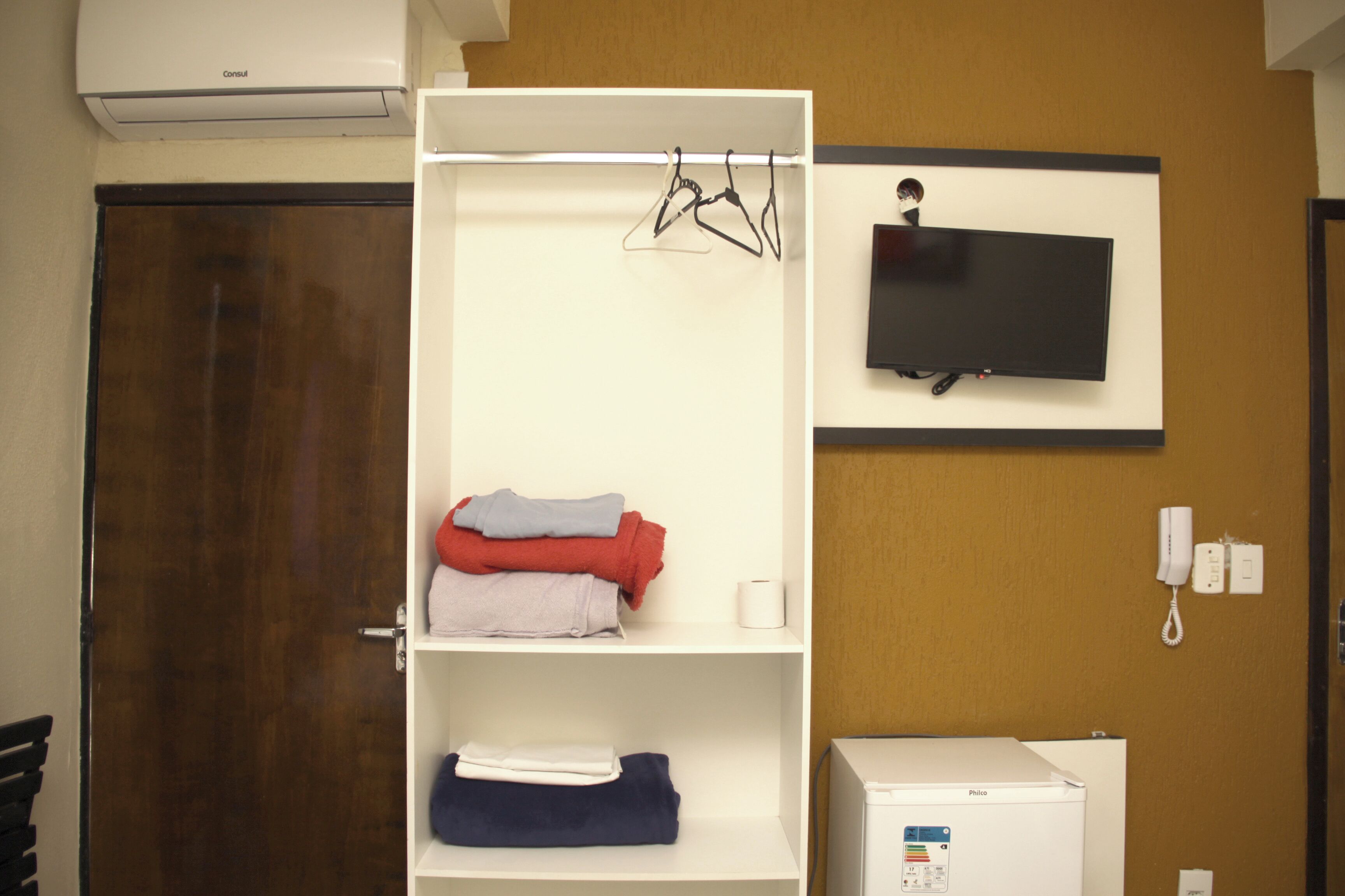 A hotel room with a white wardrobe, a flat-screen TV mounted on the wall, a bedside phone, and a small bedside table with a white appliance.