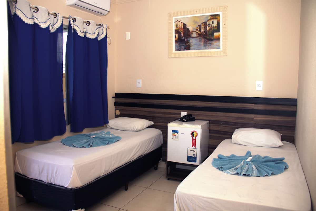Une chambre d’hôtel avec deux lits simples, une petite table de chevet avec un téléphone et un petit réfrigérateur, et un tableau accroché au mur.
