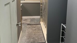 Un pasillo estrecho con una puerta a la izquierda, una ventana con persianas, una zona de ducha con puerta de cristal y un perchero metálico a la derecha.