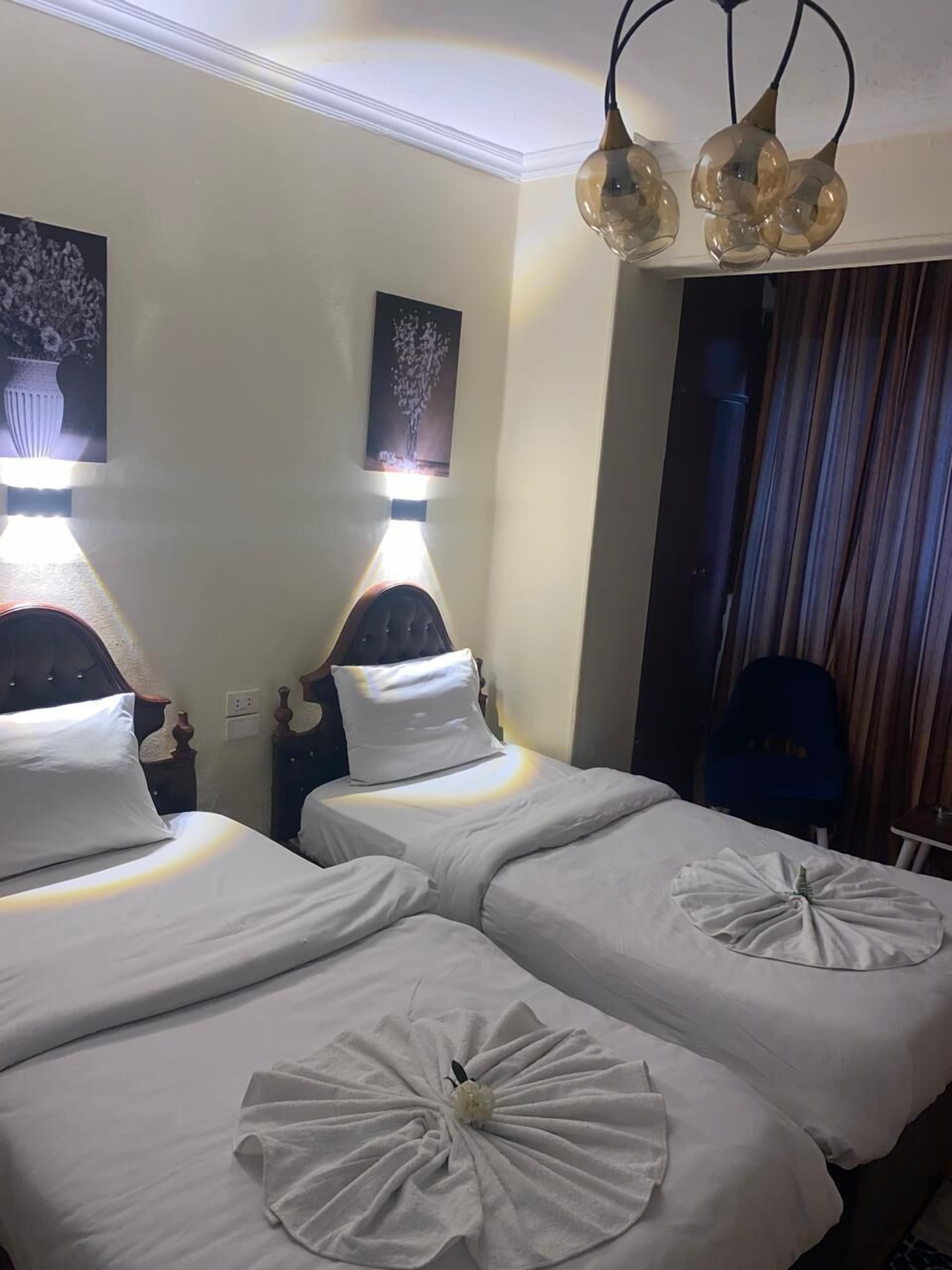 A hotel room with two beds, each with a white bedspread and a decorative towel centerpiece. There are wall-mounted light fixtures above the beds, and a dark-colored chair is visible in the corner.