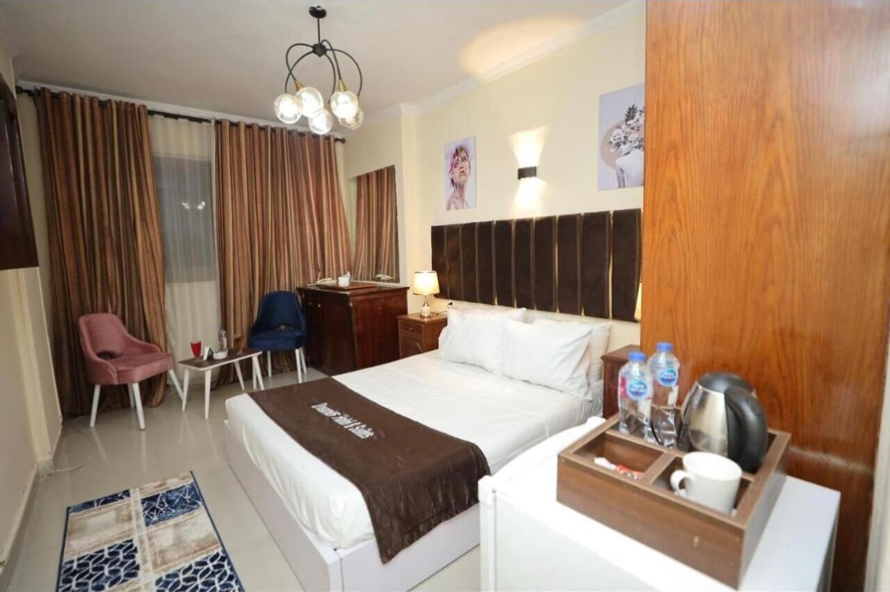 A hotel room with a bed, a desk, a chair, a nightstand, a lamp, a coffee maker, and a tray with a mug and water bottles.