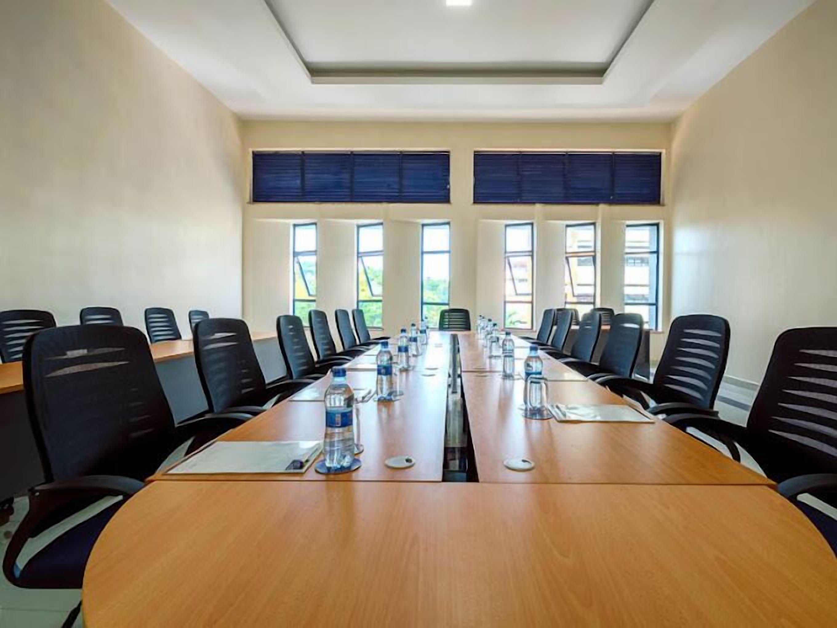 Une salle de conférence avec une longue table, des chaises noires et des fenêtres équipées de stores.