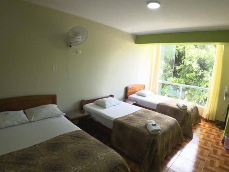 A hotel room with three beds, a window with a view of greenery, and a fan on the wall.